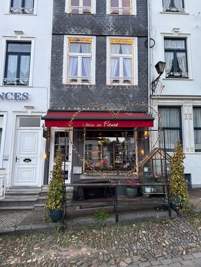 The image shows a storefront in the city of Stavelot, Belgium. The store is located at coordinates 50.395, 5.929. The store is called "L'Atelier des Fleurs" and has a red awning with a heart-shaped string of lights hanging above it. The storefront is decorated with plants and flowers. The windows are large and display a variety of floral arrangements. The store appears to be closed. To the left of the store is a white building with a white door. The door is made of wood and has a single pane of glass. The building has a black metal railing. In front of the store is a black metal bench and a stone-paved street. There is a green bush in a pot to the left of the bench.