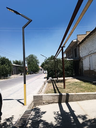 The image captures a street scene in Eshanguzar, Uzbekistan, on a clear, sunny day. A modern-looking streetlight with a zig-zag support pole stands on the left side of the frame, casting a shadow on the concrete sidewalk. The sidewalk runs alongside a paved road with a yellow and white painted line. Across the street, houses with textured walls and tiled roofs are visible, interspersed with mature trees. Two large metal pipes, one rusted and the other lighter colored, run diagonally across the upper right portion of the image, supported by visible beams. In the foreground on the right, there is a patch of dry grass and a concrete curb, with shadows of tree branches dappled across the ground. A metal grate is embedded in the sidewalk in the lower left corner. Several cars are parked on the side of the road and a few pedestrians are walking in the distance on the sidewalk. The overall scene depicts a typical urban environment in Uzbekistan during daytime.