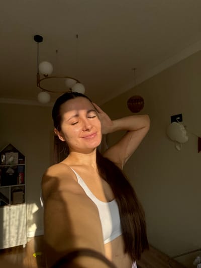 The image captures a female individual taking a selfie indoors in Dubki (Gp Kubinka), Russia. The woman, seen from the mid-torso up, has long dark hair styled in a low ponytail extending over her right shoulder. She wears a white ribbed sports bra or crop top. Her eyes are closed, and a subtle smile is present on her lips. Her left hand is positioned on the back of her head, while her right arm extends forward, holding the camera, casting a shadow across her lower chest and abdomen. Strong, direct sunlight illuminates the left side of her face and upper body, creating distinct highlights and shadows.

The background shows a room with light-colored walls and ceiling. A modern chandelier featuring a black frame and multiple white spherical lights hangs from the ceiling in the upper left. A smaller, dark spherical pendant light is also visible on the ceiling further to the right. To the left of the woman, a white, multi-compartment shelving unit, possibly shaped like a house, is visible, containing various small items. Sunlight casts pronounced shadows from this unit onto the wall and floor. To the right, a white, cloud-shaped decorative item is affixed to the wall, with a string of small, partially visible red flags or bunting hanging beneath it. Light-colored flooring is present at the bottom left.
