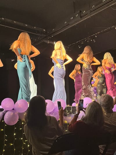 The scene depicts an indoor event on a stage in Palos Verdes Estates, United States. Approximately seven to nine women are visible on stage, predominantly viewed from the back, wearing full-length evening gowns. From left to right, prominent figures include: a woman with long brown hair in a teal backless gown with a back bow; a woman with long blonde hair in a shimmering blue-purple ruched halter gown; a woman with long dark hair in a dark metallic gown with a laced back; and a woman with blonde hair in a shimmering fuchsia-magenta backless gown. Several women on stage have their hands positioned on their hips, suggesting a posing or modeling action. The stage background is dark, with overhead structural elements featuring numbered labels (e.g., 15, 16, 17, 18, 19, 28, 29, 58, 59, 60, 61) and spotlights.

The front edge of the stage is decorated with multiple lavender-colored balloons and strings of warm white fairy lights.

In the foreground, three audience members are visible from the chest up, seated. The individual on the left, with dark hair, holds a smartphone vertically, pointed towards the stage. The central individual, with blonde hair, holds a smartphone vertically with both hands, also directed at the stage, and wears a white wristband on the left wrist. The individual on the right, with grey hair, observes the stage. The audience area is dimly lit in contrast to the illuminated stage.