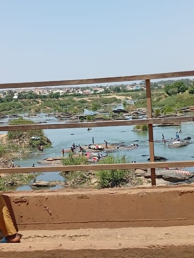 The image displays a wide view of a river in Nassarawa, Nigeria, observed from an elevated structure, likely a bridge. In the immediate foreground, the lower leg and foot of an individual wearing light trousers and a blue sandal are visible, positioned against a reddish-brown concrete barrier. A metal railing, composed of two horizontal bars and vertical posts, spans the mid-foreground, partially framing the scene.
The midground is dominated by a light blue-green river flowing around numerous dark, flat rock formations. Approximately 20-30 individuals are present on and around these rocks and in the shallow water. Many are engaged in washing activities, with various colored fabrics (red, white, blue, green) laid out on the rocks, suggesting laundry is in progress or drying. Some individuals are seen bent over, actively scrubbing, while others stand upright in the water or on the rocks.
The riverbanks feature sparse green vegetation and exposed earth. In the background, beyond the river, a sprawling settlement with numerous buildings, characterized by light-colored roofs, is visible against a clear, pale blue sky. Trees are interspersed throughout the distant urban landscape.