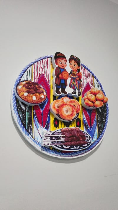 A circular decorative wall hanging, approximately 40-50 cm in diameter, is affixed to a plain light grey wall in Moynaq, Uzbekistan. The central surface displays a traditional Uzbek ikat pattern in red, blue, yellow, and white, encircled by a blue and white braided border. Applied to this surface are raised cardstock cut-outs creating a three-dimensional effect. These include two cartoon-style figures positioned centrally: a boy in a brown jacket and black skullcap holding a patterned disc, and a girl in a colorful striped dress and matching skullcap holding a small bowl containing a dish. Four additional cut-outs depicting traditional Uzbek dishes are distributed around the figures: a white bowl of plov with meat and white legumes at the upper left, a plate of four round flatbreads (non) with a central indentation below the boy, a bowl of numerous small golden-brown pastries (samsa or manty) at the upper right, and a plate of several grilled meat skewers (shashlik) at the bottom center. The assembly is evenly illuminated, casting minor shadows.