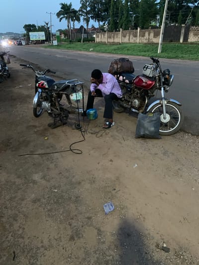 A man is repairing a motorcycle on the side of a road in Gbeleko, Nigeria. The man is wearing a light purple shirt and dark pants. Two motorcycles are visible. One motorcycle is mostly disassembled, and the man is kneeling and working on the exposed engine. The other motorcycle is mostly intact, with baggage strapped to the seat. The road is paved on one side and a dirt shoulder is on the other side. A black bag is laying near the intact motorcycle. A clear plastic wrapper is on the ground on the dirt side of the road. Trees are visible in the background. Buildings are across the road, with distant city lights in the lower left.