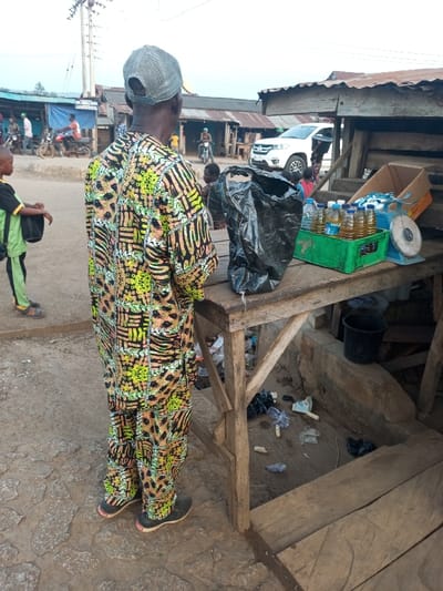 The image captures a bustling street market scene in Aramoko, Nigeria, viewed from behind a central figure.
The main subject, a person seen from the back, wears a vibrant, full-length two-piece outfit (likely a tunic and trousers) adorned with a striking pattern of black, yellow-green, and light orange geometric and organic shapes. They also wear a grey mesh baseball cap and dark sneakers with white soles. This individual stands facing a simple, crude wooden market stall.
On the stall, merchandise is displayed: a green plastic crate holds numerous small, clear plastic bottles filled with a yellowish liquid, likely cooking oil or palm oil. Beside the crate, a light blue manual scale with a round metal weighing pan sits, along with a small, open cardboard box and a large, black plastic bag. Underneath the wooden stall, the ground is dirtied with scattered litter, including plastic bags and several white corn cobs, suggesting an active, open-air environment. A black plastic bucket also sits on the ground below the stall.
In the background, the scene reveals more market activity. Several people are visible, including a child in a green and black shirt on the left. Simple, single-story market stalls or shops with corrugated metal roofs line the street. Motorcycles with riders are present, and a white SUV can be seen further down the road. Power lines and a utility pole are visible against the sky.
The time of day appears to be daytime, likely late morning or early afternoon, characterized by bright but soft natural light under an overcast or hazy sky, with no harsh shadows. The overall atmosphere is one of everyday commerce and community life in an informal market setting. No clear, readable text is visible on signs or objects in the image.