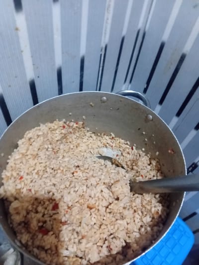 A close-up shot shows a pot filled with cooked rice mixed with small red pieces, possibly peppers. A metal spoon rests in the rice. The background is a wall with vertical grey and black stripes, and a blue object is visible at the bottom right. The scene is likely in a home kitchen in Pantinapu, Nigeria.