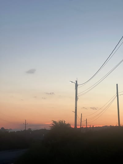 The image depicts a dusky sky over a rural landscape in Bârnova, Romania. Power lines stretch across the frame, supported by wooden utility poles. The sky transitions from a pale blue on the left to a warm gradient of orange and pink on the right, suggesting the sun is either rising or setting. A few wispy clouds are scattered across the sky. The lower portion of the image is dominated by dark, silhouetted foliage and a road. In the distance, faint lights can be seen, hinting at habitation. The overall mood is peaceful and serene, evoking the quietude of dusk in a countryside setting. There is no visible text in the image.