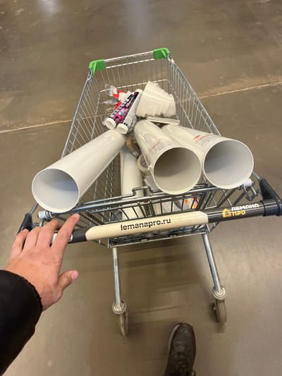 An overhead, first-person perspective image depicts a person pushing a metal shopping cart filled with building materials inside a Lemana Pro (Leroy Merlin Pro) store in Sholokhovo, Russia. The person's right hand, wearing a dark sleeve, grips the cart's light-colored handle, which displays "lemanapro.ru" and "ЛЕМАНА ПРО" (Leroy Merlin Pro) branding. The person's left foot, shod in a dark shoe, is visible on the light gray, industrial-style floor. The cart contains several large, light gray PVC pipes, multiple tubes of sealant or adhesive with white, red, and black labels, and a stack of white plastic cups or containers. Other indistinct items are also present within the cart.