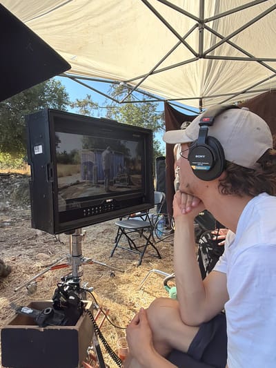 A young man wearing a baseball cap and Sony headphones is observing a video monitor on a film set, likely in the city of Mate Yehuda Regional Council, Israel. The monitor displays a scene with two portable changing rooms or shower stalls set up in an outdoor environment, possibly a desert or dry, bushy area. The man is seated, with his arms resting on his knees, and his chin is supported by his hand as he intently watches the screen.
The monitor is mounted on a tripod, and other film equipment, including a camera and a wooden case, are visible in the foreground. The set appears to be shaded by a large white canopy, supported by a metal frame, which is partially visible in the upper portion of the image. The ground is covered with dry straw or hay, with sparse vegetation and trees in the background. The overall setting suggests a production taking place outdoors, possibly for a film or television show, under bright daylight conditions.