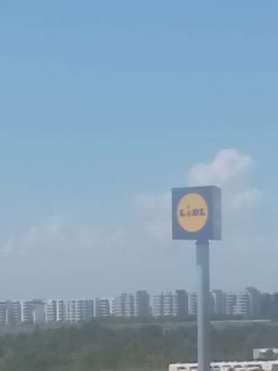 The image captures a daytime scene in Crețuleasca, Romania, dominated by a clear blue sky with scattered white clouds. In the mid-ground, a tall pole supports a square sign with a yellow circular logo displaying the text "LIDL." The sign is partially obscured by the pole and appears slightly out of focus. In the background, a strip of green vegetation is visible, beyond which a row of modern apartment buildings stretches across the horizon. These buildings are varied in height and design, with some featuring balconies and multiple floors. The overall impression is one of a suburban or semi-urban environment on a sunny day.
