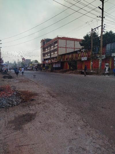 A deserted street scene under a cloudy sky. A multistory building, painted white with red trim, towers over storefronts with closed, red shutters. Power lines crisscross overhead. People walk along the distant sidewalks. A heap of gravel and orange safety fencing sits on the near left side of the road. The asphalt is cracked and worn. A dog wanders across the street.