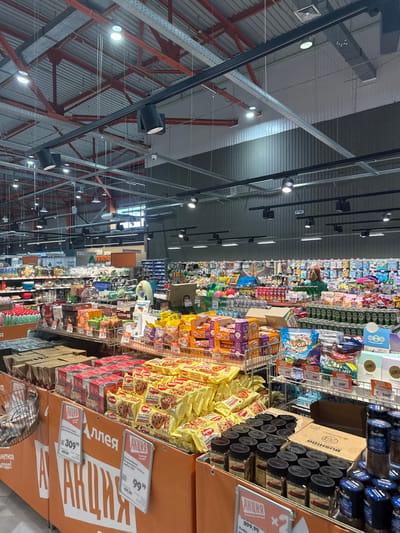 This image shows the interior of a large supermarket or hypermarket, likely in Angarsk, Russia, given the Cyrillic text. The perspective is from a high vantage point, looking down the aisles, and the image is rotated 90 degrees clockwise.
The aisles are stocked with a wide variety of consumer goods, including packaged foods like cookies, candy, and snacks, as well as beverages and other household items. There are promotional signs and displays throughout the store, indicated by the prominent "АКЦИЯ" (Aktsiya), meaning "Sale" or "Promotion" in Russian. Some of these signs feature prices like "99", "109,99", "179", "259", "309", "349", "499,99", and "899,99". Brand names visible include "LEBO", "Jardin", "BUSHIDO", "TESS", "КОРОВКА" (Korovka - a type of candy), and "Победа" (Pobeda - Victory).
The lighting appears to be artificial, typical of an indoor retail environment, with rows of spotlights visible on the ceiling structure. The architecture suggests a modern, expansive building with exposed metal beams and a corrugated metal ceiling.
There are no people clearly visible in the frame, but the presence of merchandise and promotional materials indicates that the store is open and likely has customers browsing, even if they are not in focus or easily discernible from this angle. The overall impression is of a busy commercial space stocked for shoppers.