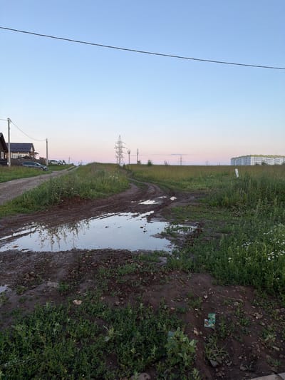 This image depicts an outdoor scene in Фролы, Russia, likely at dusk due to the soft, pastel hues in the sky and the dim lighting. The landscape features a muddy, unpaved road with large puddles, suggesting recent rainfall or poor drainage. Green vegetation, including grass and wildflowers, grows alongside the road, with some plants appearing waterlogged. In the distance, a row of tall apartment buildings is visible, hinting at a semi-urban or suburban environment. Power lines and transmission towers are also prominent features of the landscape, indicating the presence of infrastructure.
Several cars are parked along the dirt road, and a house with a dark roof is also visible. The overall mood is somewhat somber and reflective, amplified by the muddy conditions and the muted colors. No people are clearly discernible in the image, and no text is visible. The reflection of the sky and surrounding vegetation in the puddles adds a sense of depth and atmosphere to the scene. The image is oriented vertically.