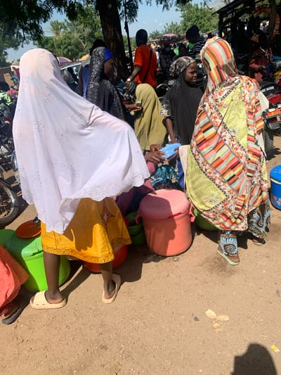 The image depicts a group of people, likely in Boi A, Nigeria, gathered around what appears to be a transportation hub or marketplace. The orientation of the image is vertical, with the subjects appearing in a somewhat jumbled and layered arrangement.

The main subjects are several individuals, primarily women, dressed in colorful and patterned clothing, including headscarves and long dresses. One woman in the foreground is wearing a white garment adorned with small, dark decorative elements. Another is wearing a vibrant, patchwork-style dress. A woman in a black garment and headscarf is visible further back.

Numerous objects are scattered amongst the people, including large plastic containers in various colors (blue, green, pink, and orange), likely used for storage or transport. Some of these containers are quite large and seem to be serving as makeshift seating. There are also what appear to be bags and possibly parts of motorbikes, suggesting this is an area where people are preparing to travel or have just arrived. One person is holding a small blue object.

The setting appears to be an outdoor, unpaved area, with sand and dirt visible on the ground. Sunlight casts distinct shadows, indicating it is daytime, likely mid-morning or afternoon given the intensity of the shadows. The presence of trees and foliage in the background suggests a rural or semi-rural location.

The activity seems to be related to travel or waiting for transportation, with people sitting on or near their belongings. The overall mood is difficult to ascertain due to the angle and cropping, but the scene appears somewhat crowded and informal. There are no discernible emotions expressed by the individuals. No visible text is present in the image.