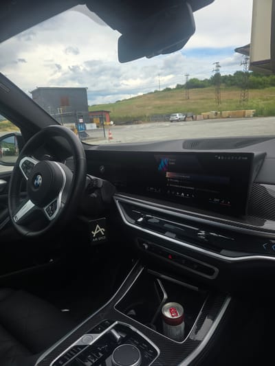 This media file presents an interior view of a modern car, likely a BMW, with a focus on the dashboard and steering wheel. The subjects are primarily the car's interior elements, including the infotainment screen, steering wheel with the BMW logo, a can of what appears to be Coca-Cola, and a carbon fiber trim. The scene is set inside the car, which is parked, as suggested by the static view and the external environment visible through the windshield.
The infotainment screen displays information such as "13:38" for the time, and messages related to the car's functions, like "Activate pre-cooling now" and "Some online services restricted." This indicates an ongoing event of the car being prepared for use or experiencing connectivity issues. The steering wheel suggests the driver's perspective, though no people are directly visible.
Notable details include the carbon fiber accents on the dashboard, adding a touch of luxury and sportiness. The presence of the Coca-Cola can hints at a casual use or a break. The external view shows a cloudy sky and a somewhat industrial or rural setting with power lines and buildings, suggesting a location like Balanovo, Bulgaria, as provided. The lighting appears to be natural daylight, possibly mid-afternoon given the time displayed. There are no strong emotional cues conveyed by the image. The visible text includes various labels on the car's controls and the infotainment system, as well as the brand name "BMW" and "AIR SPICE" on an air freshener.