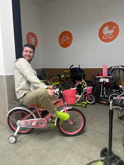 A man is sitting on a child's pink bicycle with training wheels, indoors. The man, wearing a grey hoodie and khaki pants, is smiling at the camera. The bicycle has a pink basket on the handlebars and a decorative sticker on the chain guard. The background shows a collection of other bicycles, scooters, and a stroller, suggesting a storage area or a shop. On the wall are orange circular stickers with white icons of a palette and paintbrush, a bicycle, and a baby carriage. The floor is tiled. The location is specified as Мирный, Russia.