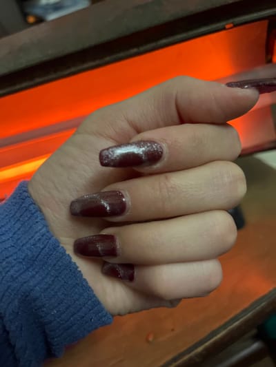 The image displays a close-up view of a person's hand, primarily focusing on their manicured nails. The hand appears to be an adult's, likely female, and is partially clad in a blue, ribbed long-sleeved garment, possibly a sweater or jacket, visible on the lower right side.

The nails are long and shaped in a square or coffin style, painted a deep, dark red or maroon color. They feature a distinct glittery or shimmering finish, which appears to have a "cat-eye" effect, creating a diagonal streak of lighter sparkle on each nail. This effect is particularly noticeable on the index, middle, and ring fingers. There's a slight gap visible between the cuticles and the start of the nail polish on some fingers, suggesting either nail growth or an application technique.

The hand is positioned above a prominent, bright orange glowing light source, which occupies a significant portion of the left and central background. This light source could be a nail drying lamp, a heater, or another form of illumination, casting an intense, warm glow upwards onto the fingers. The immediate surroundings are out of focus and dark, with hints of a dark, possibly wooden, surface to the right and above the glowing light. There is no visible text in the image. The overall lighting suggests an indoor setting with a focused light source.