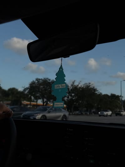 The image captures a daytime scene from within a vehicle, located in Aventura, United States. A teal, pine tree-shaped "Little Trees" air freshener, labeled "Caribbean Colada," hangs centrally from the rearview mirror support structure. The vehicle's dark dashboard and a portion of its infotainment screen, displaying "Radio" and frequency options, occupy the lower frame. A human hand, adorned with a ring on the fourth finger, is visible on the left side, resting on what appears to be a steering wheel or dashboard.
Through the windshield, an outdoor environment is visible under a blue sky with scattered white cumulus clouds. Multiple vehicles are present in the midground, including a silver sedan parked or slowly moving in the center-left, and several white SUVs/trucks further back to the right. In the background, a distinctive yellow and red Shell gas station canopy is discernible. The scene is populated by numerous mature green trees, indicative of a developed urban or suburban landscape. No explicit human figures or active interactions are directly observed beyond the hand inside the vehicle.