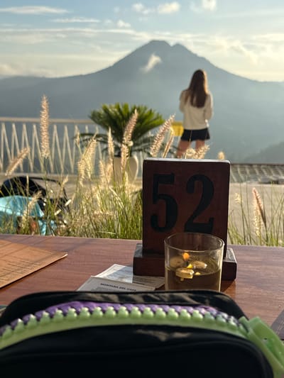 The image, taken in Kedisan, Indonesia, shows a partially obscured table at a restaurant with a view of a mountain. The table has a wooden stand with the number "52" displayed, a glass with decorative elements inside, a menu, and a black bag with purple and light green details. In the background, beyond a railing with decorative grasses, a woman wearing a white top and dark shorts is visible. A palm tree is to the left of her and in the far background is the large mountain. The sky is light and slightly overcast.
