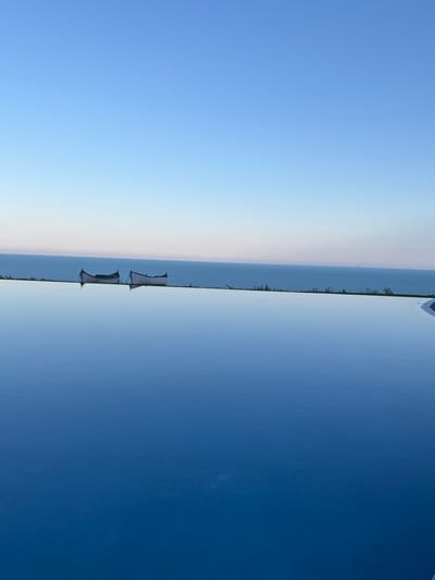 The image depicts an infinity pool overlooking the Black Sea in Topola, Bulgaria. Two small, white, wooden boats are positioned along the edge of the pool, on a strip of green vegetation separating the pool from the sea. The sky is clear and blue, transitioning to a lighter hue near the horizon. The water in the infinity pool is calm and reflects the sky and the boats. The scene conveys a sense of tranquility and scenic beauty.
