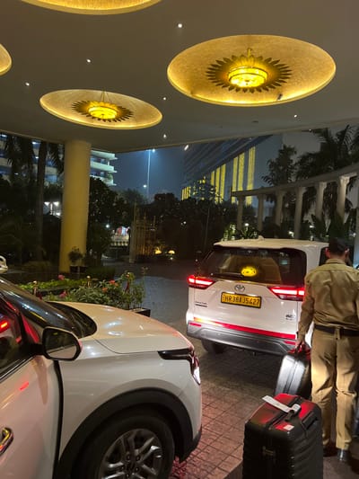 The scene depicts the well-lit porte-cochère of a modern luxury hotel or commercial complex in Noida, India, at nighttime. The overhead structure features a light-colored ceiling with three large, circular, recessed lighting fixtures, each emitting a golden glow from a central light source and adorned with a radiating sunburst pattern. A large, light-colored cylindrical support pillar is visible to the left.
In the foreground, the front right quarter of a white SUV, likely a Kia Seltos, is partially visible with its front passenger door slightly ajar. To its right and slightly behind, a white Toyota Innova Hycross SUV with taillights illuminated is parked or idling. This vehicle displays a yellow Indian license plate "HR 38 AJ 5427", indicating Haryana registration, and a circular yellow sticker with a sunburst design visible through its rear window.
To the right of the Toyota SUV, a male individual is seen from behind, wearing a light brown uniform with a dark cap. He is carrying two black hard-shell suitcases with wheels; one is held by the handle in his right hand, and the other is being pulled alongside him. The ground beneath is dark, patterned paving, showing reflective wet patches.
Landscaping elements, including small flower beds with red and pink blossoms, potted plants, and bushes, are situated between the vehicles and the main pillar. In the background, beyond the covered area, dark trees and an ornate decorative gate are visible. Further back, a multi-story building is illuminated with vertical lines of yellow lights, contributing to the urban night environment. A street light illuminates a hazy background.