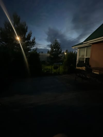 The image depicts a dimly lit outdoor scene at dusk in Bol'shoe Zaborod'e, Russia. A tall, illuminated street lamp is visible on the left, casting a strong beam of light that cuts diagonally across the frame, partially obscuring a dark evergreen tree. In the background, a corrugated metal fence stretches across the scene, with sparse vegetation and trees visible beyond it. To the right, the corner of a brick house with a green roof is visible. The sky is dark and cloudy, suggesting approaching evening or stormy weather. The ground in the foreground appears to be a wooden deck or patio, also dark and indistinct. No people or specific actions are identifiable in the image.