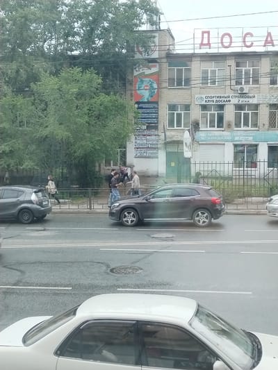 The image depicts a street scene in Chita, Russia, with a building in the background displaying Cyrillic signage, including "ДОСААФ". In the foreground, several cars are visible on the wet asphalt road. A dark gray hatchback is parked on the left side of the road, and a brown Mercedes-Benz SUV is positioned further down the street. A white sedan is partially visible at the bottom of the frame. On the sidewalk behind a metal fence, a group of four people are interacting. Two men in the center appear to be engaged in a discussion or argument, with one man in a light-colored jacket gesturing. Another man in a dark jacket is standing slightly behind them, and a fourth person in a light-colored jacket and dark pants is walking away from the group. Trees with green foliage are situated behind the fence, and overhead wires crisscross the scene. The overall lighting suggests an overcast day.