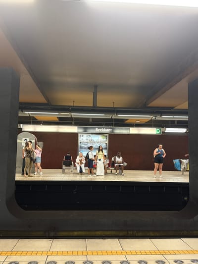 The image shows a subway platform in Beekant, Molenbeek-Saint-Jean, Belgium. Several people are visible, some standing and some sitting on benches. The platform is clean and well-lit, with a maroon-colored wall behind the benches. There is a sign that reads "Beekant" above a large advertisement. In the foreground, the edge of the platform with tactile paving is visible. The overall atmosphere is one of a typical urban public transport hub.