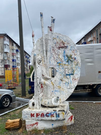 The image shows an outdoor sculpture in what appears to be a residential area of Usolye-Sibirskoye, Russia. The sculpture is primarily white with multi-colored paint splatters (red, blue, yellow) and features a disc-like shape with protruding extensions. It is positioned on a base covered in similar white material and paint. "KREACHOK" is written on the base in red paint, which is likely a misspelling of "Крючок", meaning "Hook" in Russian.
Behind the sculpture are residential buildings and a playground, suggesting a public space. A white commercial truck is partially visible behind the sculpture. The sky is overcast. A car and a wooden log are also visible in the foreground.