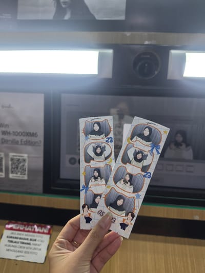 This media file captures a close-up view of a person's hand holding two photo strips from a photo booth. The photo strips feature stylized illustrations of two women, seemingly friends, posing in a playful manner. One strip has the numbers "23" and "053" printed on it, possibly indicating a date or a sequence number. The background reveals a photo booth, with its reflective surface showing another image of the women. A sign on the lower right corner in red and yellow reads "PERHATIAN" (Attention) followed by instructions in Indonesian, which seem to be related to photo quality. Below that, some text indicates "Win WH-1000XM6 Danilla Edition?" suggesting a promotional event. The scene appears to be indoors, possibly at a mall or entertainment venue in Medan, Indonesia, as suggested by the location context. The lighting is bright and artificial, typical of such locations. The overall mood is casual and fun, reflecting the enjoyment of taking commemorative photos.