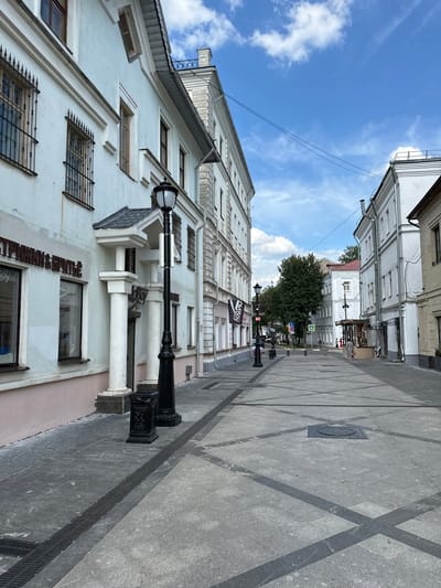 The image shows a street scene in Putyatino (Semivragovskaya S/A), Russia, on a partly cloudy day. The perspective is from a low angle, looking down a pedestrianized street lined with buildings.
On the left, a pale blue building features barred windows on the upper floors and a pink-painted lower section. The signage on the building is in Cyrillic and appears to advertise a barber shop or hair salon. A black lamppost with an ornate base stands prominently in the foreground. White columns support an overhang above an entrance to this building.
The street itself is paved with light gray stone tiles arranged in a pattern of intersecting lines, with darker gray diagonal stripes forming a grid. Grates are visible along the edges of the street.
On the right, a white building with multiple windows stretches down the street. Further down, other buildings of varying architectural styles and colors are visible, including a red-roofed building. A few people are present in the distance, walking along the street and near an outdoor seating area. Trees are visible further down the street, with green foliage. The sky above is blue with scattered white clouds.