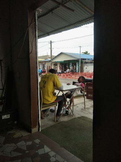 The image depicts an outdoor scene in Cotonou, Benin, viewed from a shaded interior doorway. In the immediate foreground, a tiled floor transitioning to green artificial turf is visible. The dark wooden or metal frame of the doorway occupies the left and right edges, with exposed electrical wiring running along the left frame.

In the midground, two individuals are seated at a white, foldable rectangular table with metal legs, positioned on a concrete or dirt surface under a corrugated metal roof. An adult, seen from the back, wears a yellow t-shirt with a dark graphic and dark trousers, sitting barefoot on a chair with a green fabric seat and metal frame. A pair of black slide sandals with white stripes rests on the ground near their feet. Opposite them, a child, also barefoot, is reclined in a similar chair, wearing a light-colored (possibly pink) t-shirt and light shorts. The child looks upwards and to the left with an open mouth, appearing to be laughing or speaking. A pair of black and white striped slide sandals are near the child's feet. To the right of the child, a stack of brown crates, labeled "SCHILDERIA" and "SUBETRA," is present.

The background reveals a street or open area undergoing what appears to be construction, indicated by numerous reddish-orange plastic safety barriers. Utility poles with overhead power lines are visible. Several buildings line the street: a cream-colored structure with a prominent green-trimmed storefront displaying merchandise, and a larger, lighter-colored building with a blue corrugated roof further in the distance. A person on a blue scooter or motorcycle is partially visible moving along the street on the far left. The sky is overcast and bright.