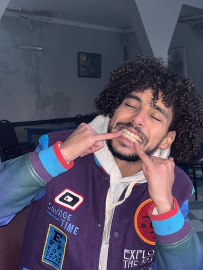 A man with curly dark hair and a beard is smiling, pointing to his teeth with two fingers. He is wearing a hoodie with a varsity jacket. The jacket is purple with patches on the sleeves. The sleeves are a mix of purple, blue and green. The man is indoors and the room has poor lighting. There is a small framed picture on the wall in the background.