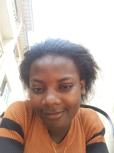 A close-up, slightly upward-angled portrait of a young adult female, taken outdoors in Agburu Uke, Nigeria, is depicted. The subject has dark skin, black curly hair, and wears an orange-brown ribbed long-sleeved top with two black horizontal stripes on the visible left arm. Small, light-colored stud earrings are present on her right ear, and a thin gold-colored necklace with a pendant is visible around her neck. She is looking downwards and slightly to her left, exhibiting a subtle smile with partially closed eyes. In the upper left background, a portion of a light-colored building with dark-framed windows and an overhanging roof structure is visible. The upper right and central background consist of an overexposed bright white area, indicative of the sky. A dark-colored chair back is partially visible on the far right edge of the frame. The scene is brightly illuminated, consistent with daytime conditions.