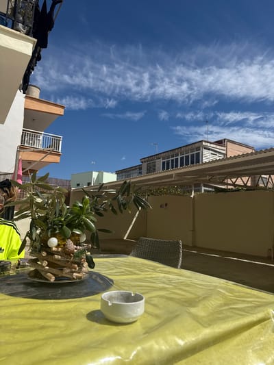 The image shows a view from a balcony or rooftop in Les Meravelles, Spain, evidenced by the architecture and sky. A table covered with a yellow tablecloth is in the foreground. On the table, there is a decorative centerpiece with twigs, leaves, and white ornaments, alongside a white ashtray. A person wearing a neon yellow and black Adidas tracksuit is partially visible near the bottom of the frame. The background features buildings with balconies and varying architectural styles under a blue sky with streaky white clouds.