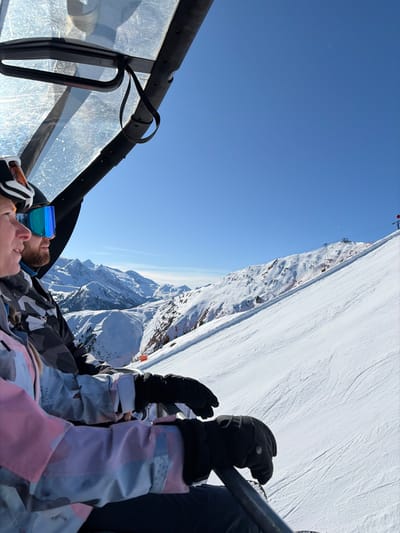 Two individuals are depicted riding an overhead ski lift in Schwendau, Austria, under a clear blue sky over snow-covered mountains. The occupant on the left, wearing a white helmet, blue mirrored ski goggles, and a pink, grey, and white patterned ski jacket, sits beside a bearded male occupant in a dark helmet, blue mirrored ski goggles, and a dark grey patterned ski jacket. Both are looking forward and to the right, observing the expansive snowy peaks and a groomed ski slope descending to their right. Their hands, clad in black gloves, rest on the ski lift's safety bar. A translucent overhead canopy of the ski lift is partially visible in the upper left of the frame.