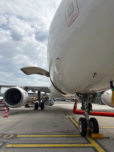 This image shows the underside of an aircraft, likely a passenger jet, at Schiphol Airport in the Netherlands. The prominent features include the plane's white fuselage, the landing gear with its wheels chocked, and one of the engines. The underside of the wing has an access panel open, and markings indicating the location of the static port. The background shows airport tarmac with yellow lines, other aircraft, and ground service equipment. The sky is visible with a partly cloudy weather.