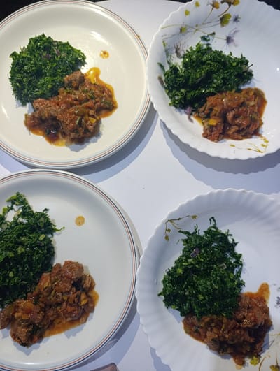 Four plates hold portions of food. Each plate contains a serving of dark green leafy vegetables and a stew-like dish. The plates are predominantly white, with two having floral patterns around the edges and two featuring simple red and blue concentric circles. The food is arranged side-by-side on each plate, with the stew appearing brownish-red in color. The entire composition is set against a bright, white surface.
