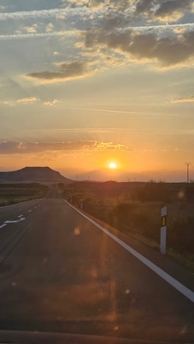 This is a vertical, wide-angle, outdoor photograph taken during a sunset. The sun, a bright orange orb, is setting behind a low, dark mountain range on the right side of the frame, casting a warm, golden glow across the sky. The sky is filled with wispy clouds, some tinged with orange and pink from the setting sun, while others are still a soft blue. Streaks of white, likely contrails from airplanes, are visible in the upper left quadrant of the image.

A paved road with a white line down the center dominates the right half of the image, curving away into the distance. The road appears to be empty, with no vehicles or people visible. On the right side of the road, there are several vertical posts with yellow and white reflective markers. Beyond the road, the landscape is hilly and covered in dry, golden-brown vegetation, suggesting a warm climate. In the distance, silhouetted against the sunset, are several tall, thin structures that appear to be power line pylons.

The overall mood of the image is serene and peaceful, with the vibrant colors of the sunset creating a sense of warmth and beauty. The location appears to be rural, given the open landscape and lack of buildings. The time of day is clearly sunset, indicated by the low position of the sun and the dramatic lighting. There is no visible text in the image.