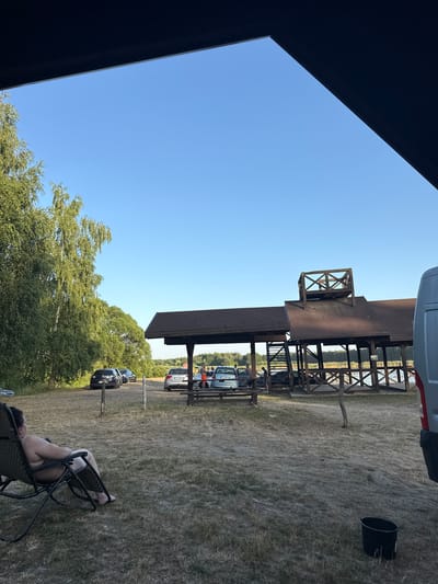 A person reclines in a chair in a grassy, outdoor setting. The scene appears to be during the daytime, with clear blue skies visible. In the background, there is a wooden structure that resembles a covered pavilion or dock, with several cars parked underneath and nearby. Trees form a backdrop to the scene, and a body of water is partially visible beyond the wooden structure. The overall atmosphere seems relaxed, suggesting a leisurely day outdoors.