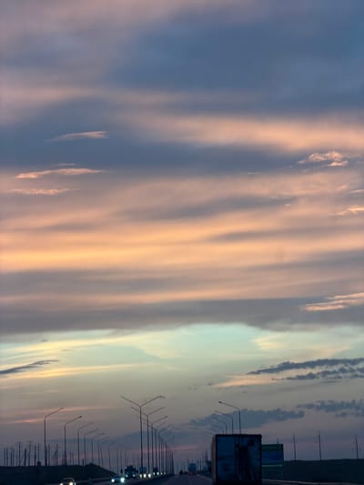 The image shows a highway at sunset in a rural area, likely near a transportation hub in Kazakhstan, given the prominent billboard for Astana in the image. The sky is filled with streaks of pink and orange clouds against a deep blue background. A long line of streetlights stretches into the distance along the highway, with a few cars and trucks visible on the road. The overall impression is one of a peaceful journey through an expansive landscape as day turns into night.