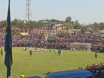A vibrant football match is captured from an elevated perspective in Siaya, Kenya. The scene depicts a large, enthusiastic crowd filling the stadium stands and surrounding areas, indicating a well-attended event. The match is in progress on a green grass pitch under clear, bright sunny skies, suggesting daytime.
On the field, football players are actively engaged in play. One team wears white jerseys with black trim, while the other is dressed in green with black trim. Several players are gathered near a goalpost, with the goalkeeper alert inside the goal, hinting at a moment of intense play or a set-piece. Various individuals in green or khaki uniforms, possibly security personnel or officials, are visible on the periphery of the field and within the crowd.
In the foreground to the left, a large flag, partially obscured, displays prominent blue, yellow, green, and red stripes. Below the viewer, a blue-roofed structure, likely a dugout or covered seating area, is partially visible. The background features a tall communications tower standing against the sky, alongside a mix of residential and commercial buildings, some appearing to be under construction or in various states of development, characteristic of an urban or semi-urban landscape. The overall atmosphere conveys a lively community sporting event. No discernible text is visible on player jerseys, banners, or signs.