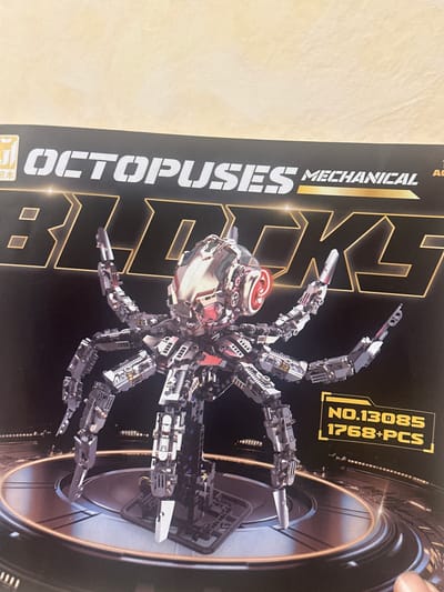 The image shows the box of a mechanical octopus building set. The box features a close-up of the assembled model, which is a metallic octopus with red glowing eyes and multiple articulated limbs. The text on the box reads "OCTOPUSES MECHANICAL BLACKS" and "NO.13085 1768+PCS". The background of the box is dark with metallic accents and a circular platform on which the octopus is standing. There is no information in the image that indicates a location in Vapniarka, Ukraine.