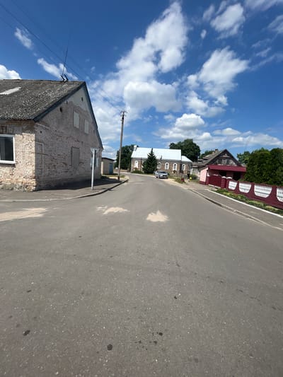 This media file depicts a street scene in Lunna, Belarus. The perspective is from ground level, looking down a paved road. The sky is a vibrant blue with scattered white clouds, suggesting a clear, sunny day, likely midday given the bright illumination.
On the left side of the road, several buildings are visible. The closest is a dilapidated brick structure with boarded-up windows, giving the impression of disuse or ongoing renovation. Further down the street, a white building with arched windows stands next to a larger, older house with a red roof and a decorative red fence. A dark-colored car is parked in front of one of these houses. Trees and greenery are interspersed with the buildings. A utility pole with wires stretches across the frame, connecting to the buildings and disappearing into the sky.
On the right side of the road, the asphalt is textured and shows some dark spots, possibly oil stains. The road curves gently out of view. The overall impression is of a quiet, rural or small-town setting in Belarus on a pleasant day. There is no discernible activity or event taking place, and no visible text.