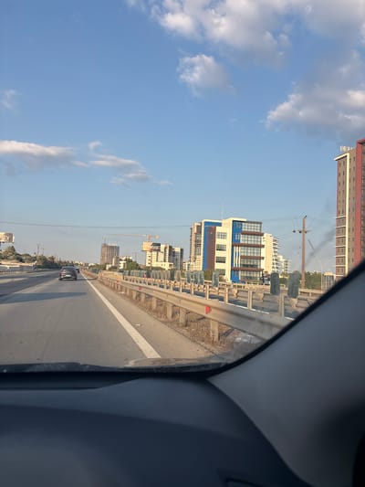A forward-facing, eye-level, exterior shot captures a view from within a vehicle looking out onto a highway. The sky is a clear, pale blue with scattered white clouds. The highway, paved with asphalt, is divided by a white line and stretches into the distance. On the right side of the highway, a metal guardrail separates the road from dry, grassy terrain. Several modern, multi-story buildings with distinctive architecture and glass facades line the roadside, some appearing to be under construction with cranes visible in the background. Billboards with text like "keon.cy" and "ency" are also present. The overall scene suggests a suburban or developing urban environment in a sunny climate.