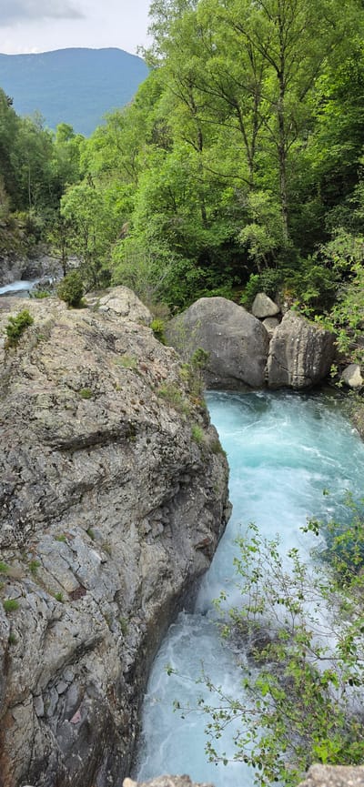 A dynamic natural landscape in Sahún, Spain, captured from an elevated perspective looking down into a river gorge. The central feature is a rapidly flowing river, characterized by its striking turquoise-blue water, which appears foamy white in turbulent areas. Large, rugged grey and brown rock formations and boulders line the riverbanks and form a prominent cliff face in the foreground.
Lush, dense green deciduous trees and various smaller shrubs cover the right bank of the river and extend across the distant mountain slopes. A large, verdant mountain rises in the background on the left side. The scene depicts the natural flow of the river through its rocky environment.
The time of day appears to be daytime, with bright, diffused lighting suggesting an overcast sky or soft sunlight. There are no human subjects or visible text. The powerful current, vibrant water color, and mountainous, forested setting are notable features of this natural environment in the Spanish Pyrenees.