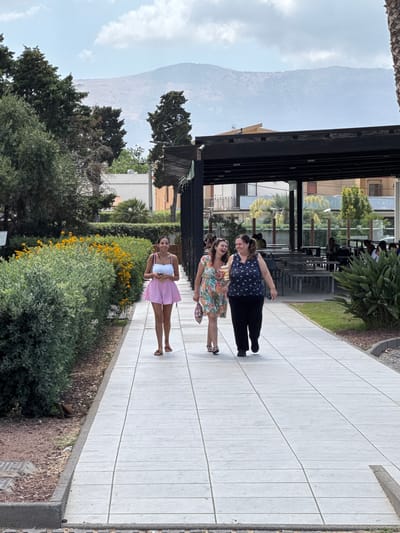 The image depicts three women walking along a paved pathway. The woman in the foreground is wearing a pink skirt and a white top. The woman in the middle is wearing a floral-patterned dress. The woman in the background is wearing a dark blue top with white polka dots and black pants. They are walking on a bright, sunlit day, indicated by the strong shadows. The pathway appears to be in a public area, possibly a park or promenade, with lush green bushes and trees lining one side. In the background, there are buildings and a distant mountain, suggesting an urban or semi-urban setting. The overall mood seems relaxed and casual, as the women are smiling and appear to be enjoying their walk. There is no visible text in the image.