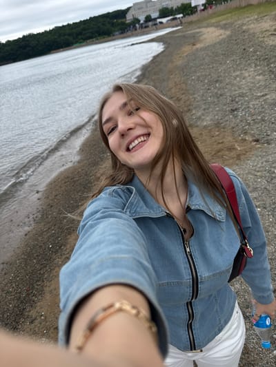 The image shows a young woman taking a selfie on the pebble beach of Russky Island, Russia. She is wearing a blue denim jacket, white pants, and a red purse over her shoulder. The water of the bay is visible to her left, and a forested hill with buildings in the distance is to the left of the bay. She is holding a bottle of water in her right hand.
