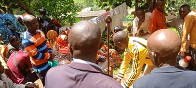A group of people gathers outdoors. Some sit or kneel, wearing colorful, patterned clothing, while others stand. At least two men in suits are visible from the back. A man in a yellow tracksuit bends forward. In the background, a clothesline with hanging clothes is seen, and a building with a corrugated metal roof peeks through the trees. A tall wooden staff with a circular ornament stands out amidst the crowd.