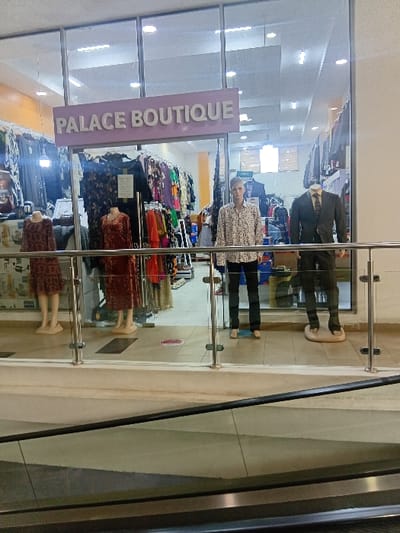 The image shows the storefront of Palace Boutique, located in the city of Pridelands, Kenya. The storefront is visible through a large glass window, showcasing a variety of clothing items displayed on mannequins. Three mannequins are visible, two dressed in women's clothing and one in men's clothing.  There is one person standing inside the shop. The person, a young man wearing a light-colored patterned shirt and dark pants, is likely a customer browsing the selection. The glass window has a metal railing in front of it, which is also visible in the foreground.  The floor in front of the storefront is tiled, and there is an escalator leading up to the next floor. The escalator is made of metal and has a glass railing.
