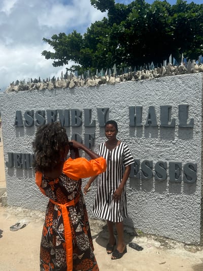 Two young women stand in front of a textured grey concrete wall with large, raised grey letters spelling "ASSEMBLY HALL OF JEHOVAH'S WITNESSES." The woman on the left, wearing a black and orange patterned dress with puffed sleeves and an orange belt, is turned away from the camera and appears to be holding a phone up to take a picture of the woman on the right. The woman on the right stands facing forward, looking towards the camera, and wears a black and white vertically striped dress and black wedge sandals. Behind the wall, lush green foliage is visible, and the top of the wall is lined with broken glass shards embedded in cement, likely for security. The ground in front of the wall is a mixture of concrete and dirt, with some debris scattered about. The sky above is partly cloudy. The location is identified as Itoga, Nigeria.